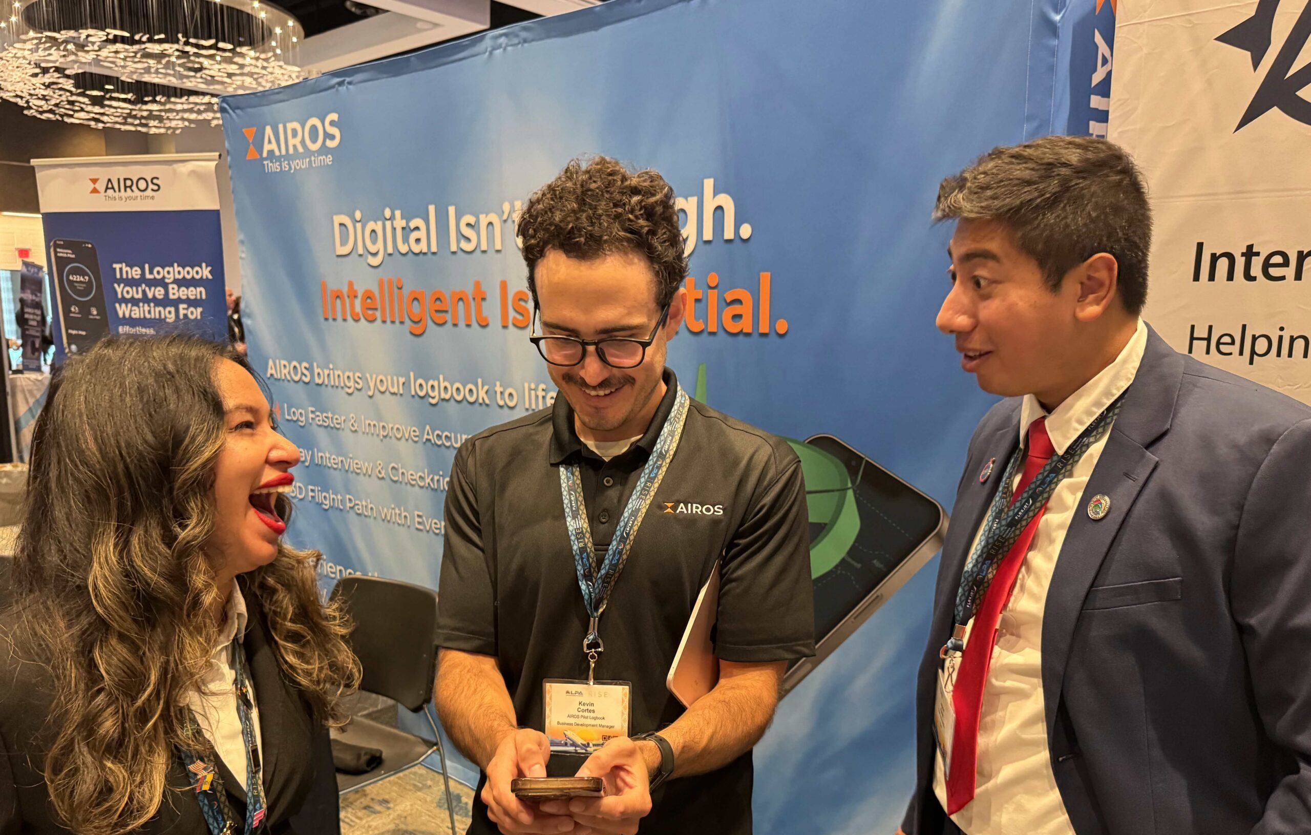 AIROS Business Development Manager Kevin Cortes shows the AIROS Pilot Logbook app on his phone to two smiling conference attendees in front of the AIROS booth backdrop reading "Digital Isn't Enough. Intelligent Is Essential."